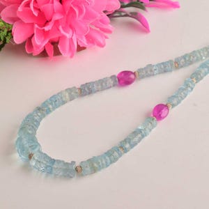 May include: A delicate necklace featuring light blue aquamarine beads interspersed with vibrant pink gemstones. The necklace is accented with small, gold-colored beads and is displayed against a white background with pink flowers.