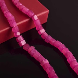 May include: Two strands of faceted, hot pink gemstone beads. The cube-shaped beads are strung together, forming a necklace or bracelet. The background is a dark, reflective surface with a red box.