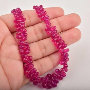 Könnte beinhalten: Eine Halskette aus facettierten, tropfenförmigen Rubinperlen. Die Perlen haben eine tiefe, lebendige rosa-rote Farbe und sind zu einer Halskette aufgereiht. Die Halskette wird in einer Hand gehalten.