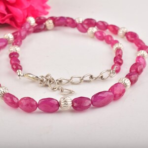 May include: A necklace with oval, deep pink gemstones and silver-coloured metal accents. The necklace is secured with a silver-coloured clasp and chain. A pink flower is visible in the background.
