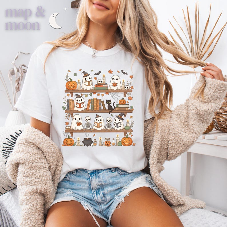 May include: White t-shirt with a Halloween-themed illustration. The design shows a bookshelf with ghosts, pumpkins, books, and a black cat. The text "map & moon" is at the top left of the shirt.