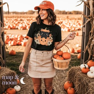 May include: A black t-shirt with a pumpkin and black cat graphic, paired with a tan skirt and orange cap. A wooden basket holds several small pumpkins. The background features a pumpkin patch.