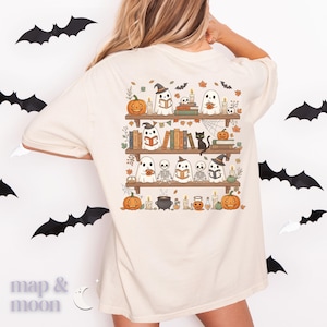 May include: Cream-coloured t-shirt with a Halloween graphic. The design shows a bookshelf with ghosts, pumpkins, books, and a black cat. Bats are scattered around the shirt. The text "map & moon" is at the bottom left.