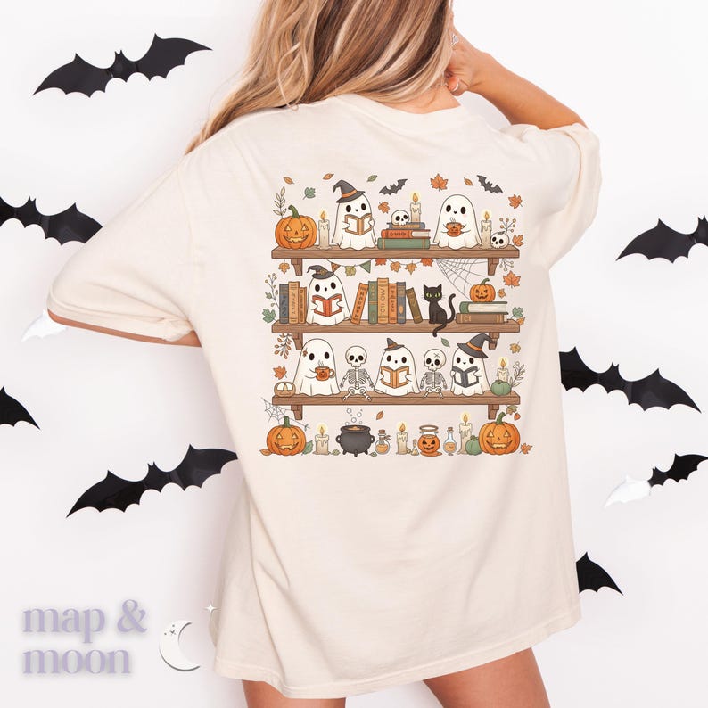 May include: Cream-coloured t-shirt with a Halloween graphic. The design shows a bookshelf with ghosts, pumpkins, books, and a black cat. Bats are scattered around the shirt. The text "map & moon" is at the bottom left.