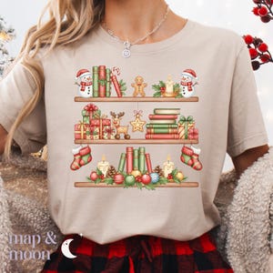 May include: Beige t-shirt with a festive Christmas bookshelf design. The graphic features three shelves filled with books, gifts, snowmen, gingerbread men, candles, and ornaments. The text "map & moon" is at the bottom.