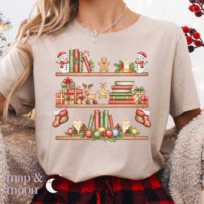 May include: Beige t-shirt with a festive Christmas bookshelf design. The graphic features three shelves filled with books, gifts, snowmen, gingerbread men, candles, and ornaments. The text "map & moon" is at the bottom.