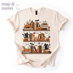 May include: Cream-colored t-shirt featuring a Halloween-themed bookshelf design. The design includes stacks of books, pumpkins, ghosts, black cats, candles, and bats. The text "map & moon" is in the upper left corner.