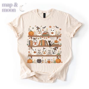 May include: A cream-coloured t-shirt with a Halloween-themed design. The graphic shows ghosts, pumpkins, books, and a black cat on bookshelves. The text "map & moon" is in the upper left corner. The design is in orange, black, and white.