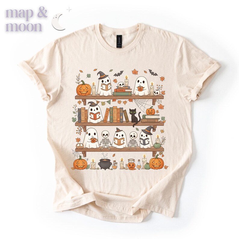 May include: A cream-coloured t-shirt with a Halloween-themed design. The graphic shows ghosts, pumpkins, books, and a black cat on bookshelves. The text "map & moon" is in the upper left corner. The design is in orange, black, and white.