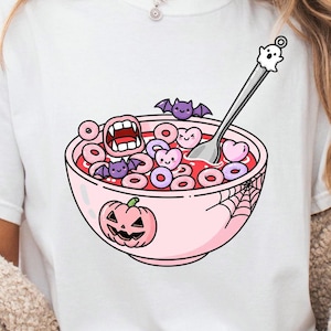 May include: White t-shirt featuring a pink bowl of cereal with Halloween-themed elements. The cereal includes heart and ring shapes, with a spoon topped with a ghost. The bowl has a jack-o'-lantern and spiderweb design.