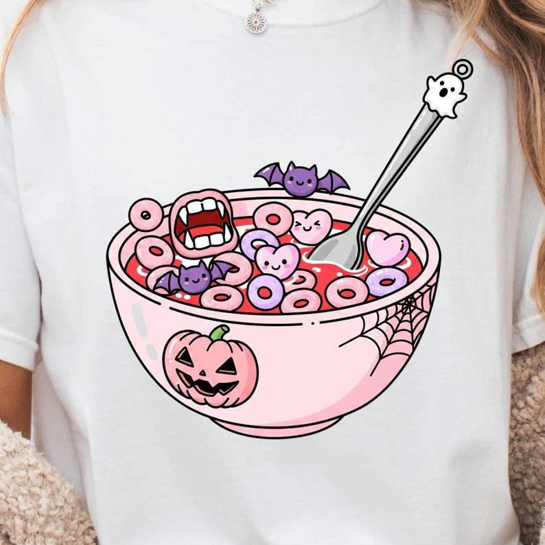 May include: White t-shirt featuring a pink bowl of cereal with Halloween-themed elements. The cereal includes heart and ring shapes, with a spoon topped with a ghost. The bowl has a jack-o'-lantern and spiderweb design.