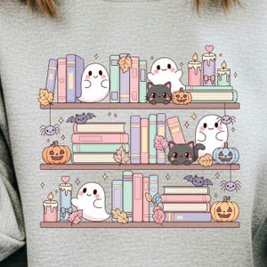 May include: A light gray sweatshirt featuring a whimsical illustration of three shelves filled with books, ghosts, black cats, pumpkins, candles, bats, and spiders. The books are in pastel colors, creating a cute Halloween theme.