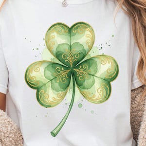 May include: White t-shirt featuring a watercolor-style shamrock design. The shamrock is a vibrant green with gold filigree accents and a green stem. The design is centered on the shirt, with subtle green paint splatters around the shamrock.