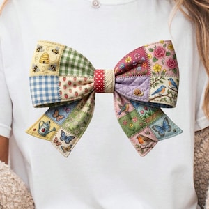 Peut inclure: T-shirt blanc avec un grand nœud coloré. Le nœud est fait de tissu patchwork avec divers motifs, notamment des abeilles, des fraises, des papillons et des oiseaux. Le centre du nœud présente un motif à pois rouges.
