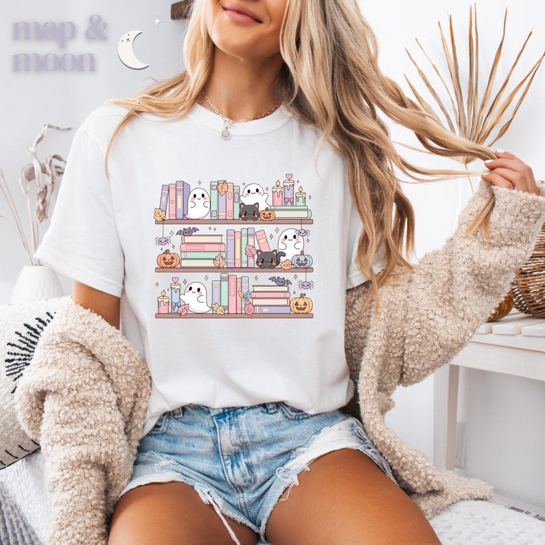 May include: White t-shirt featuring a whimsical illustration of bookshelves with books, ghosts, black cats, pumpkins, candles, and bats. The text "map & moon" is in the upper left corner. The design has a pastel color palette.