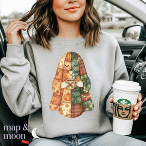 May include: A light gray sweatshirt featuring a patchwork ghost design with pumpkins, plaid, and holly. The ghost holds a mug with a festive design. The words "map & moon" are printed at the bottom left.