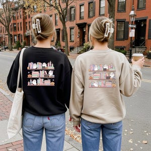 May include: Two sweatshirts, one black and one tan, feature a Halloween-themed design. The design shows a bookshelf with ghosts, pumpkins, and books. The sweatshirts are paired with blue jeans.