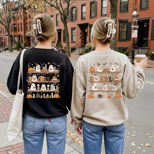 May include: Two sweatshirts, one black and one beige, display a Halloween-themed design. The design features a bookshelf with ghost characters, pumpkins, books, and other spooky elements. The sweatshirts are worn by two people.