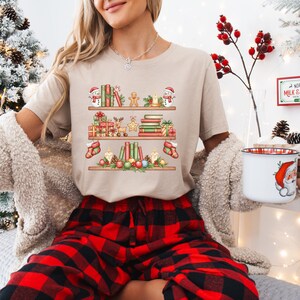May include: Beige t-shirt featuring a festive bookshelf design with Christmas-themed elements. The graphic includes snowmen, books, gifts, candles, and stockings. The model is wearing red and black plaid pajama pants.