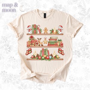 May include: Cream-colored t-shirt featuring a festive bookshelf design. The shelves are adorned with Christmas decorations, including snowmen, books, gifts, candles, and ornaments. The text "map & moon" is in the upper left corner.