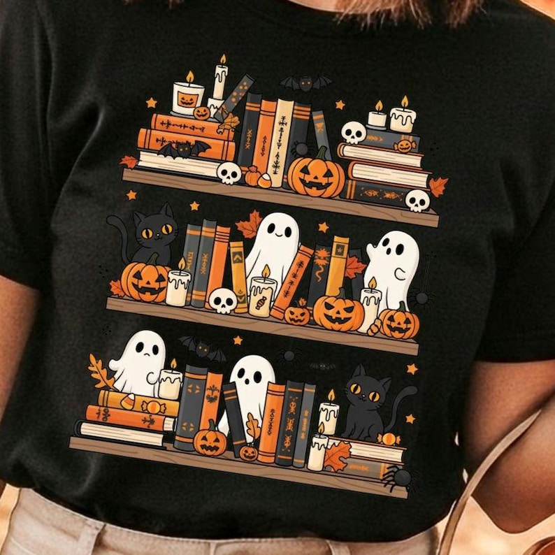 May include: Black t-shirt featuring a Halloween-themed bookshelf design. The design includes stacks of books, pumpkins, ghosts, candles, bats, cats, and skulls. The books are in shades of orange, black, and brown.