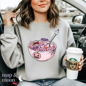 May include: A light gray sweatshirt featuring a Halloween-themed cereal bowl graphic. The bowl is pink and filled with cereal shaped like hearts and rings, with a spoon topped with a ghost. The design includes bats, a jack-o'-lantern, and a spiderweb.