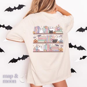 May include: Beige t-shirt featuring a whimsical Halloween-themed design. The graphic depicts three shelves of books, adorned with ghosts, black cats, pumpkins, candles, and bats. The design is in soft pastel colors, creating a cute and festive aesthetic.