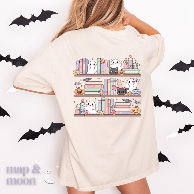 May include: Beige t-shirt featuring a whimsical Halloween-themed design. The graphic depicts three shelves of books, adorned with ghosts, black cats, pumpkins, candles, and bats. The design is in soft pastel colors, creating a cute and festive aesthetic.
