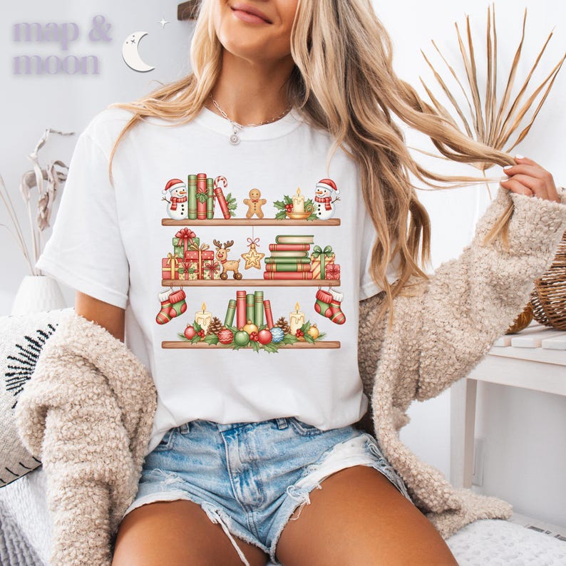 May include: White t-shirt featuring a festive bookshelf design with Christmas decorations. The design includes books, gifts, snowmen, candles, and stockings. The text "map & moon" is at the top left.