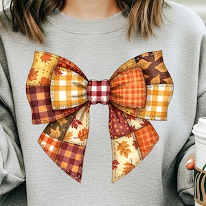 Pode incluir: Camisola cinzenta com um grande design de laço. O laço é composto por vários padrões de patchwork em tons de laranja, amarelo, castanho e vermelho, com detalhes de folhas, criando um tema outonal.