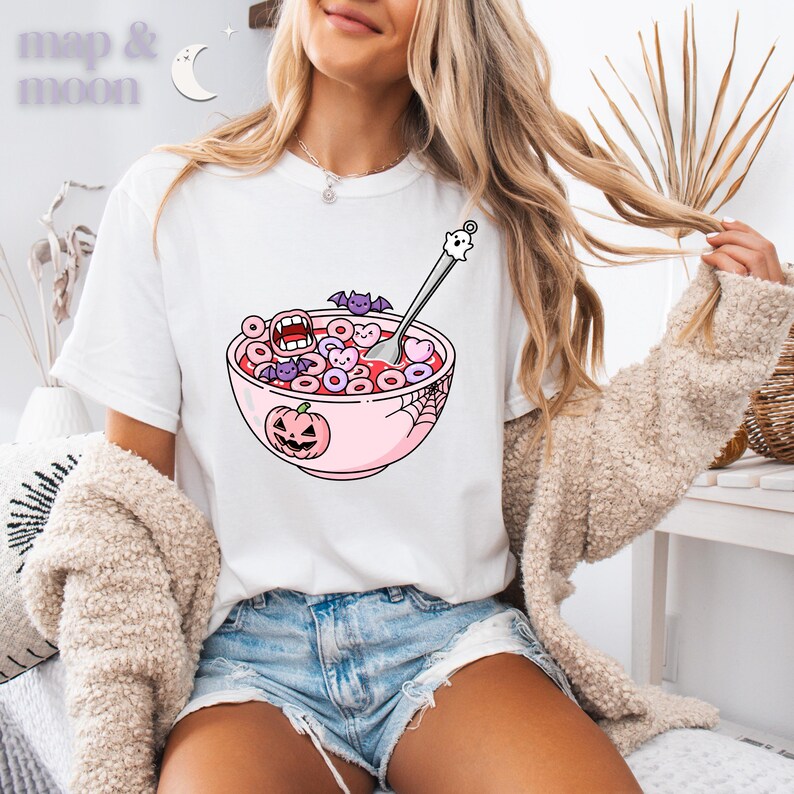 May include: White t-shirt featuring a cartoon bowl of cereal with Halloween-themed elements. The bowl is pink and has a jack-o'-lantern, bats, and a spiderweb design. The cereal pieces are heart and donut-shaped. The text "map & moon" is at the top left.
