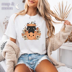 May include: White t-shirt featuring a cartoon black cat inside a pumpkin, surrounded by sunflowers and autumn leaves. The shirt has the text "map & moon" in the upper left corner. The design is in shades of orange, brown, and cream.