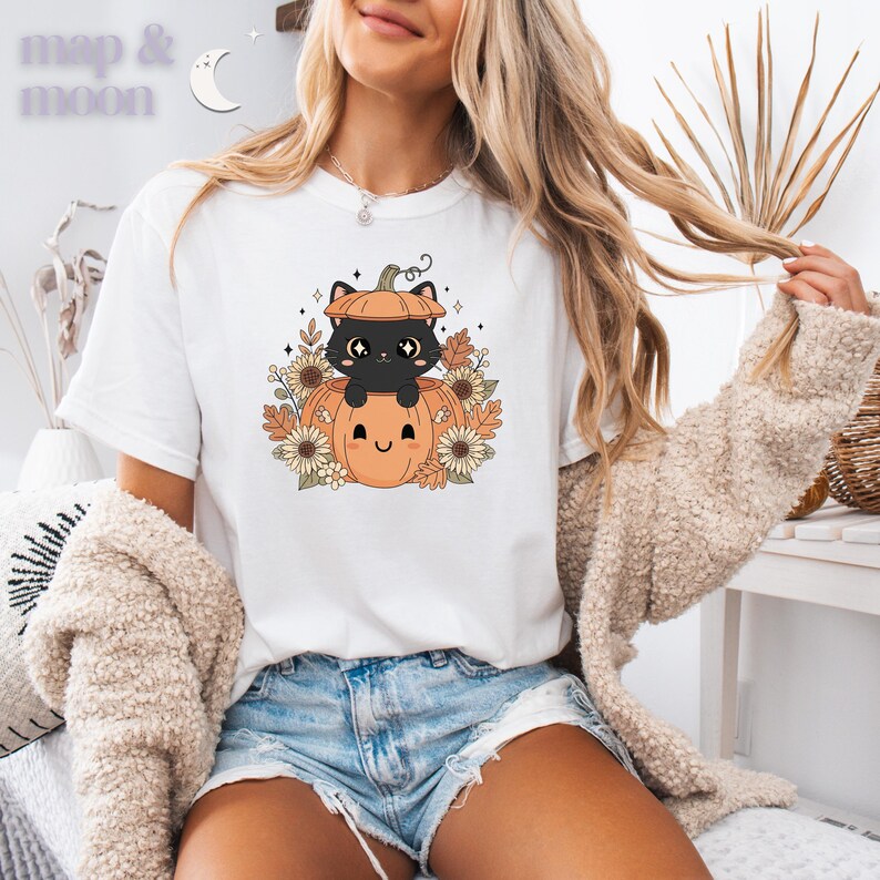 May include: White t-shirt featuring a cartoon black cat inside a pumpkin, surrounded by sunflowers and autumn leaves. The shirt has the text "map & moon" in the upper left corner. The design is in shades of orange, brown, and cream.