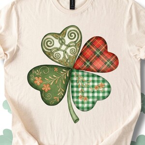 Op de afbeelding: Crèmekleurig T-shirt met een klavertjevier-ontwerp. Elk hartvormig blad heeft een uniek patroon: Keltische knopen, rood geruit, groen ruitjespatroon en bloemendetails. De klaver is groen, met een groene stengel.