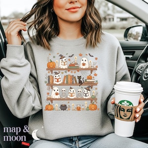 May include: A grey sweatshirt with a Halloween design. The image shows bookshelves with ghosts, pumpkins, candles, and books. The text "map & moon" is at the bottom left of the sweatshirt.