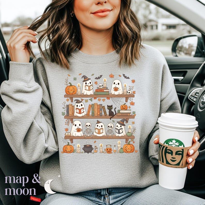 May include: A grey sweatshirt with a Halloween design. The image shows bookshelves with ghosts, pumpkins, candles, and books. The text "map & moon" is at the bottom left of the sweatshirt.