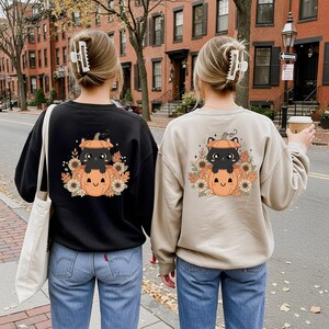May include: Two sweatshirts, one black and one beige, feature a Halloween-themed graphic on the back. The design includes a black cat sitting in a pumpkin, surrounded by sunflowers and fall foliage. The sweatshirts are paired with blue jeans.