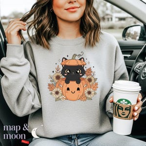 May include: A light gray sweatshirt featuring a cartoon design of a black cat inside an orange pumpkin. The pumpkin is surrounded by fall foliage and sunflowers. The text "map & moon" is at the bottom left.