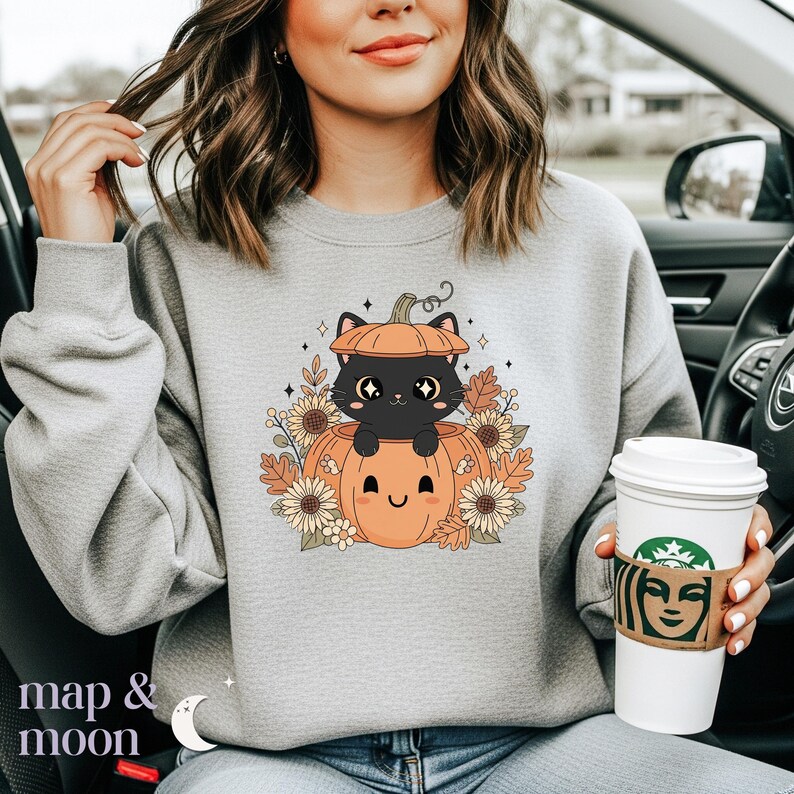 May include: A light gray sweatshirt featuring a cartoon design of a black cat inside an orange pumpkin. The pumpkin is surrounded by fall foliage and sunflowers. The text "map & moon" is at the bottom left.