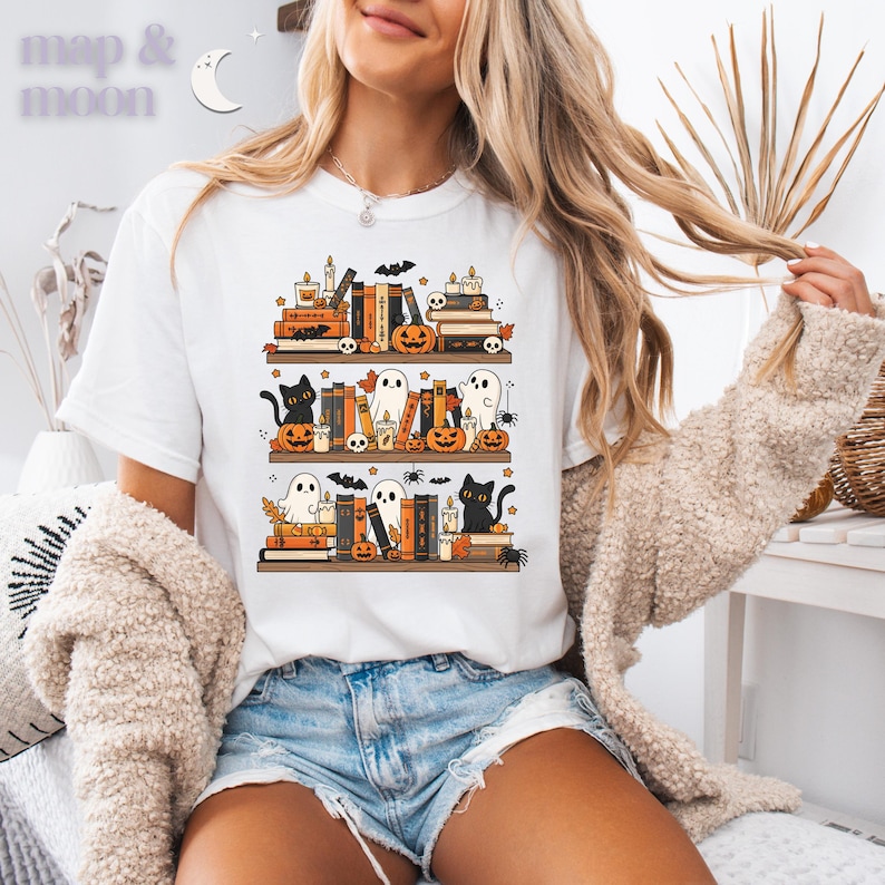 May include: White t-shirt featuring a Halloween-themed design. The graphic depicts three shelves of books, pumpkins, ghosts, black cats, candles, and bats. The text "map & moon" is in the upper left corner.