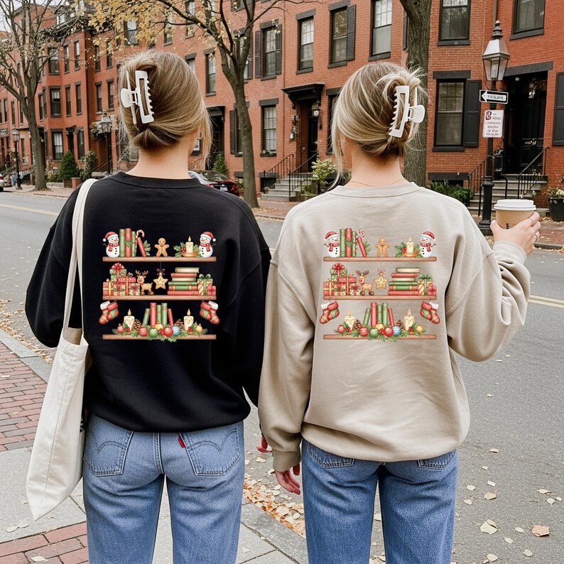May include: Two people wearing sweatshirts with a Christmas bookshelf design on the back. One sweatshirt is black, the other is tan. The design includes books, gifts, snowmen, and other holiday decorations.