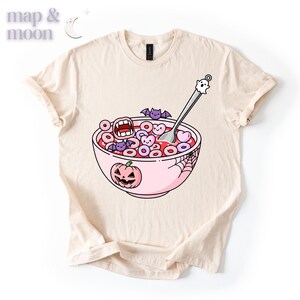 May include: Beige t-shirt featuring a whimsical Halloween-themed graphic. The design includes a pink bowl of cereal with spooky shapes, a jack-o'-lantern, bats, and a ghost spoon. The shirt has a classic crew neck and short sleeves.