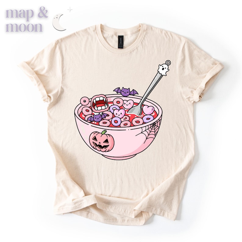 May include: Beige t-shirt featuring a whimsical Halloween-themed graphic. The design includes a pink bowl of cereal with spooky shapes, a jack-o'-lantern, bats, and a ghost spoon. The shirt has a classic crew neck and short sleeves.