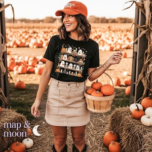 May include: Black t-shirt with a Halloween graphic of ghosts, pumpkins, and books. The person wears a rust-coloured cap with "Pumpkin Spice". A basket of pumpkins is held. The background shows a pumpkin patch.