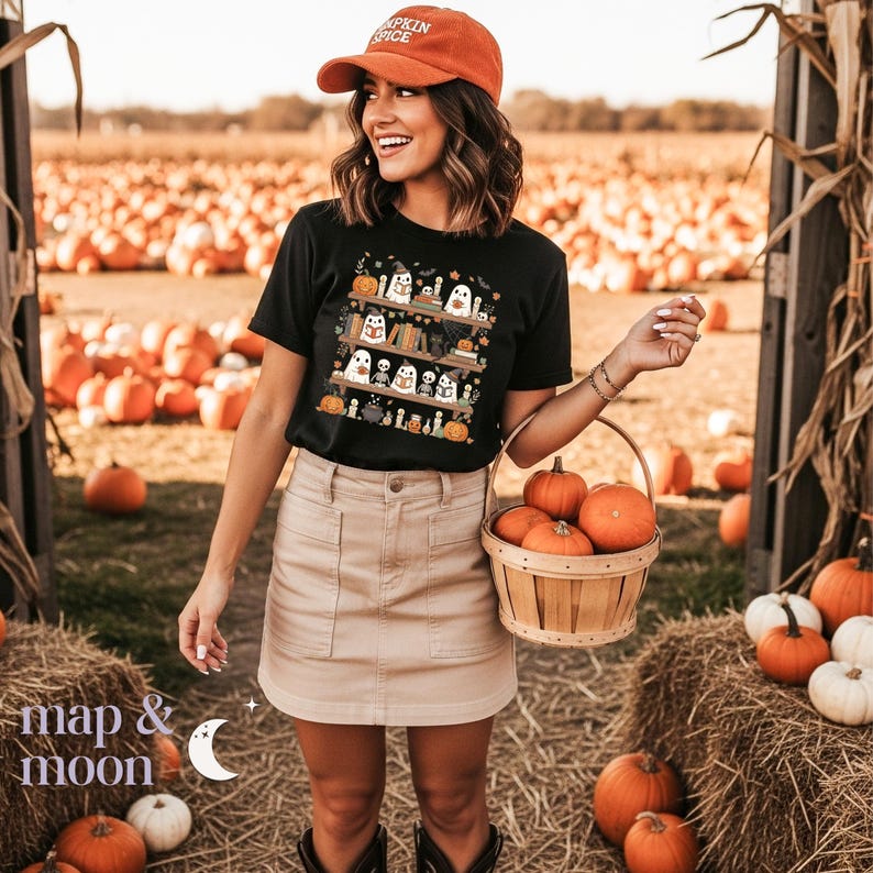 May include: Black t-shirt with a Halloween graphic of ghosts, pumpkins, and books. The person wears a rust-coloured cap with "Pumpkin Spice". A basket of pumpkins is held. The background shows a pumpkin patch.