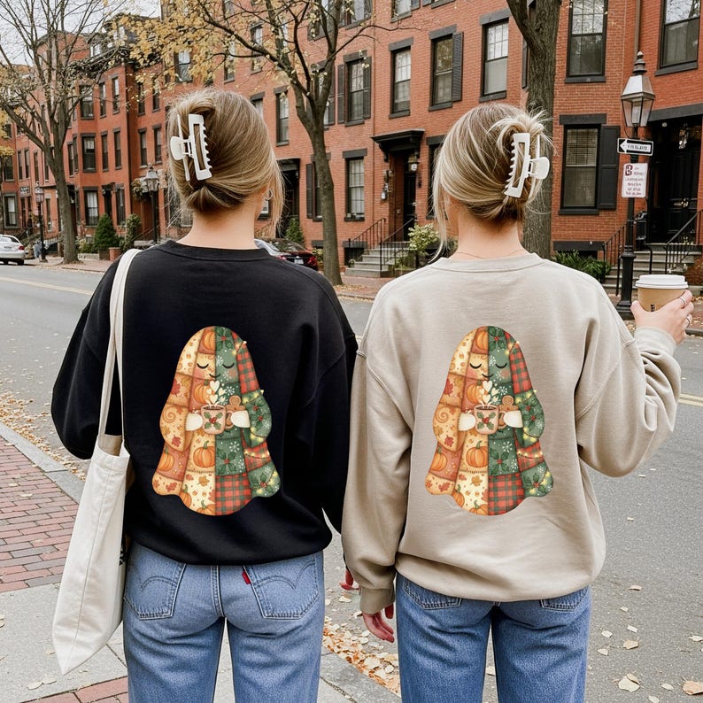 May include: Two sweatshirts, one black and one tan, each with a colorful graphic on the back. The graphic is a patchwork design with pumpkins, leaves, and a figure holding a mug. The wearers are standing on a sidewalk.