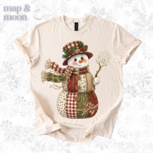 以下が含まれることがあります： クリーム色のTシャツ。雪だるまのデザイン。雪だるまはチェック柄の帽子とスカーフを身に着け、雪の結晶を持っています。デザインには赤、緑、ベージュのパッチワーク模様が組み込まれており、人参の鼻とボタンのディテールがあります。「map & moon」のブランド名が左上にあります。