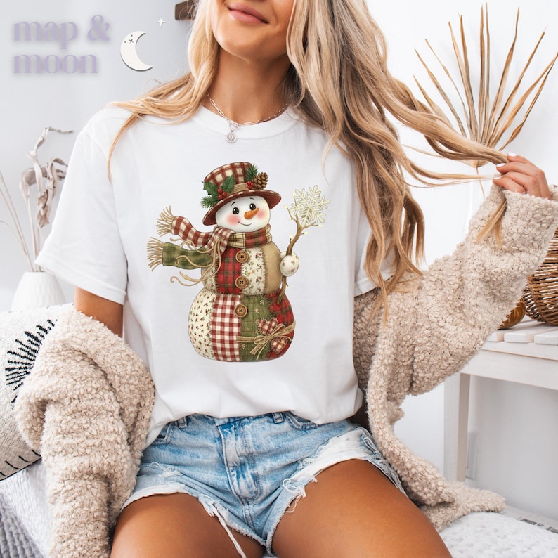 以下が含まれることがあります： 雪だるまのデザインが施された白いTシャツ。雪だるまはチェック柄の帽子とスカーフを身に着け、雪の結晶を持っています。シャツの左上に「map & moon」の文字があります。