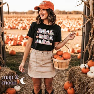 May include: A black t-shirt with a graphic of bookshelves, ghosts, and pumpkins. The person in the image is wearing an orange cap with the text "Pumpkin Spice". A basket of pumpkins is held in the person's hand. The background is a pumpkin patch.