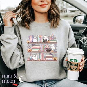May include: A light gray sweatshirt featuring a whimsical bookshelf design with ghosts, black cats, pumpkins, books, candles, and autumn leaves. The text "map & moon" is at the bottom left.